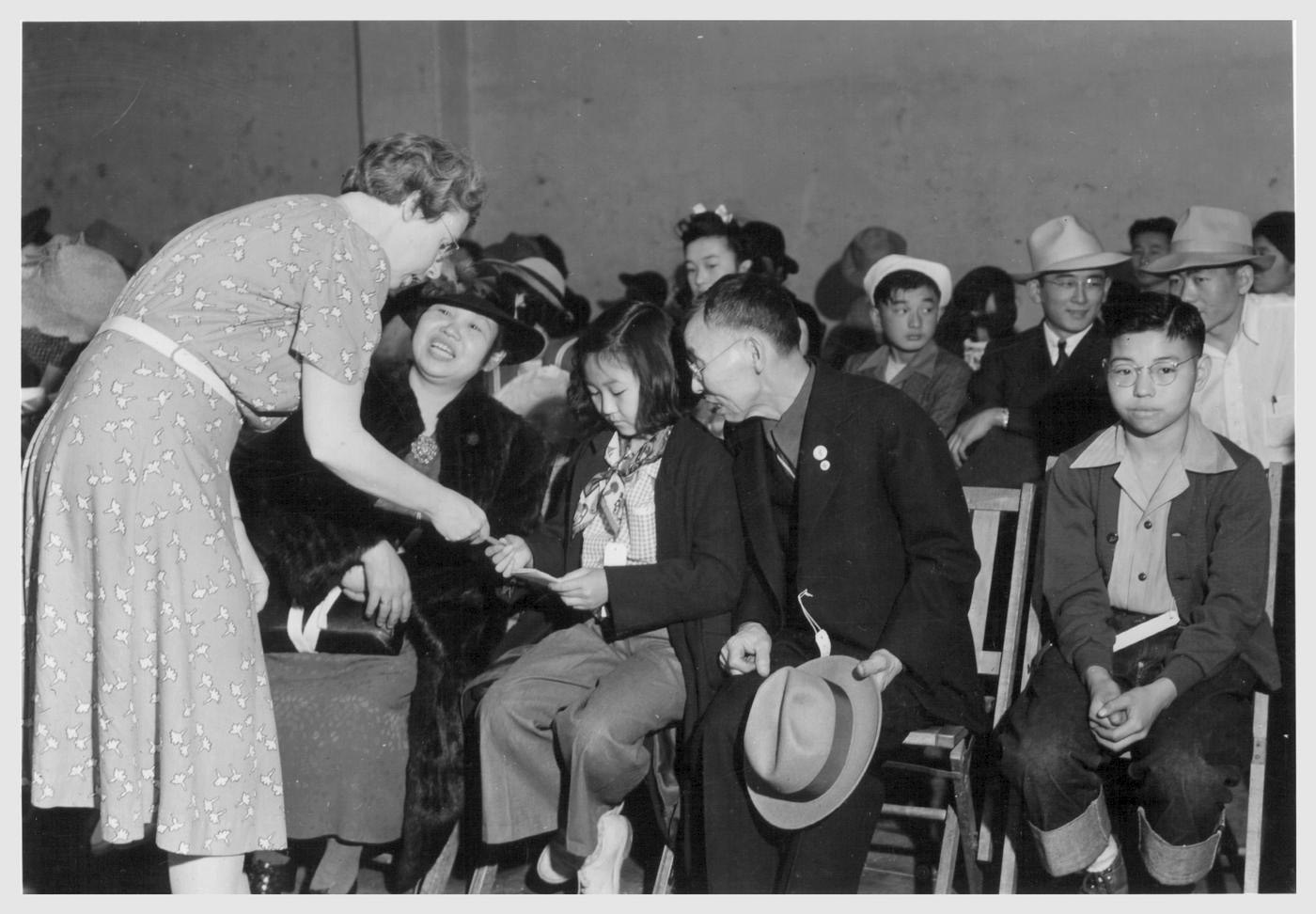 Paul Kitagaki Jr.Õs family waits to depart from 1117 Oak Street, the W.C.C.A. (Wartime Civil Control Authority) Control Station, in Oakland in 1942 for the Tanforan Assembly Center. KitagakiÕs grandmother, Juki Kitagaki, 53, is seated at left. Kimiko, his aunt, then 11, receives a pamphlet from a family friend, Dorothy Hightower, expressing her churchÕs good wishes. Grandfather Suyematsu Kitagaki, 65, watches. The photographerÕs father, Kiyoshi, 14, is at right. Family number 20247 Photographer: Lange, Dorothea -- Oakland, California. 5/ 6/ 42 Original Caption: Oakland, Calif.--Mr. and Mrs. Kitagaki with two of their children at the WCCA Control Station a few minutes before departure by bus for Tanforan Assembly Center. A local church member is handing Kimiko Kitagaki a pamphlet expressing the good wishes of the church toward the departing evacuees. Mr. Kitagaki, prior to evacuation, was in the cleaning and dyeing business. -- Photographer: Lange, Dorothea -- Oakland, California. 5/6/42 Identifier: Volume 62 Identifier: Section G Identifier: WRA no. C-581 Collection: War Relocation Authority Photographs of Japanese-American Evacuation and Resettlement Series 14: Preevacuation