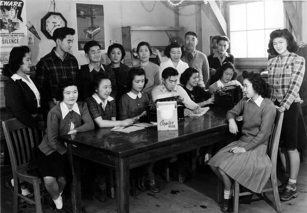 The staff of the Minidoka Irrigator, the newspaper of the Minidoka concentration camp, is shown in the paper's office. Far right (left to right): Takako Matsumoto (left) and Elsie Sata. Front (left to right): unidentified, Kimi Tambara, Cherry Tanaka, John Kanda, Miyuki Inouye, and Sachi Yasui. Back: Mitsuko Miyoshi, Harry Nakata, Watson Asaba, unidentified, unidentified, Mitsu Yasuda, unidentified, and (first name unknown) Iwami. The paper was allotted a small office space in one of the barracks that housed camp inmates. Kinoshita Collection. 