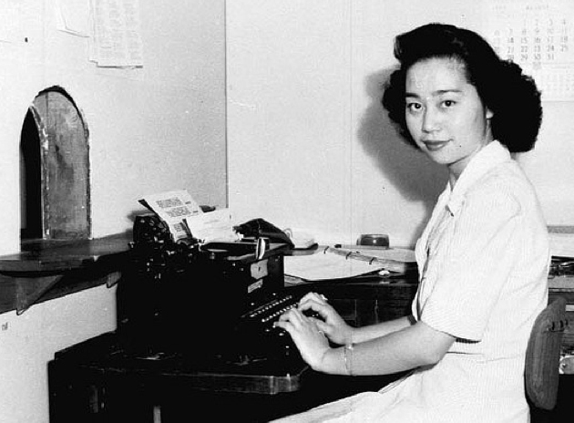 Mitsuye Endo sitting at a desk typing on a typewriter. She is looking at the camera with a slight smile.