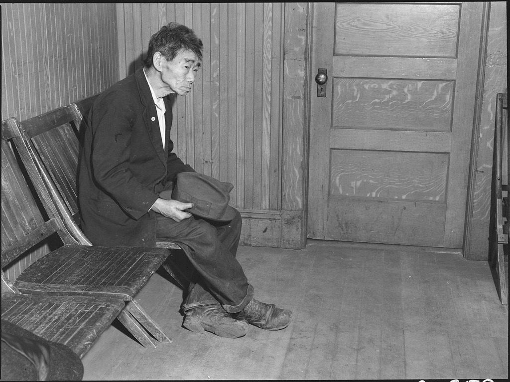 Original caption: Byron, California. Toshi Mizoguchi waiting at the Wartime Civil Control Administration station to register for evacuation. Mr. Mizoguchi came to the United States from Japan in 1892 and has been a farm laborer on California ranches since that time. He is unmarried. He is seen wearing an American flag on a celluloid button.