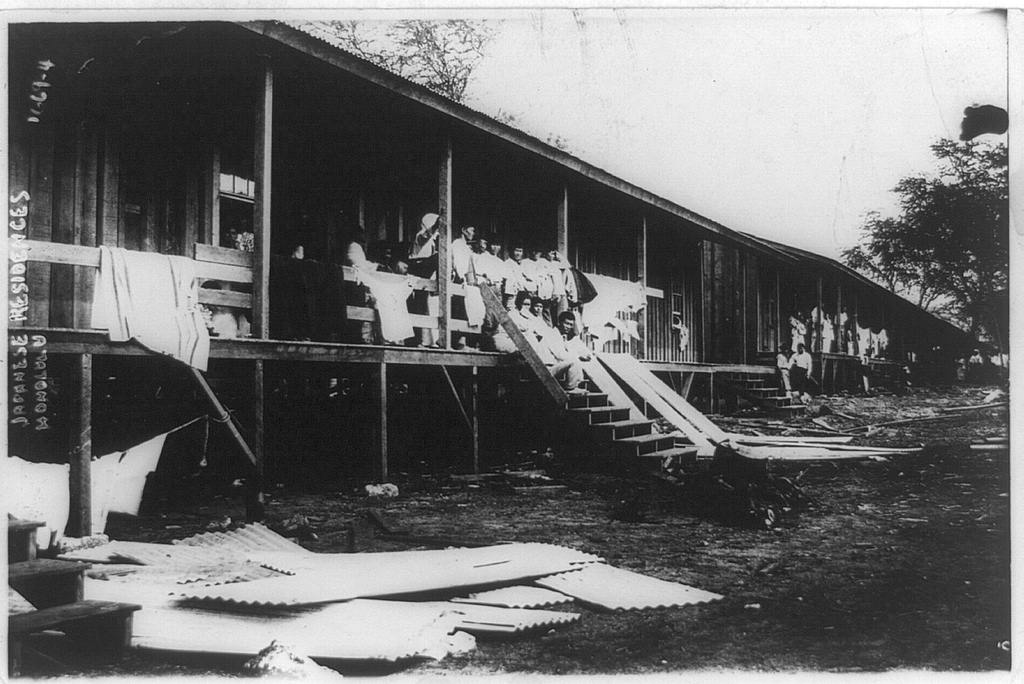 Housing for Japanese workers and their families on a plantation in Honolulu circa 1900 to 1915. Workers and their families are sitting and standing in front of the housing units. Clothing and bedding are hung up to dry, and a pile of what appears to be corrugated metal is on the ground.
