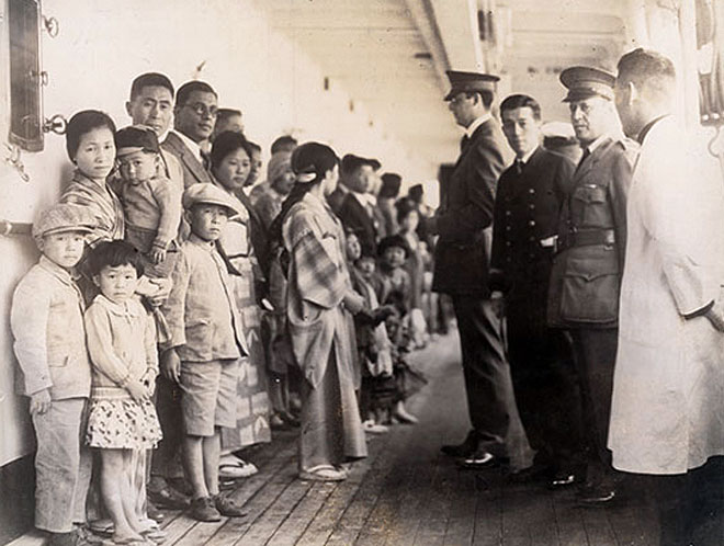 Black and white photo of Japanese immigrants standing opposite immigration officials aboard a ship. They are waiting to be examined for entry to the U.S. while docked at Angel Island.