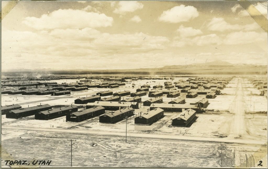 An aerial view of Topaz concentration camp, with rows of black military barracks between dirt roads and mountains in the distance. There are swirls of dust visible in the air over the camp.