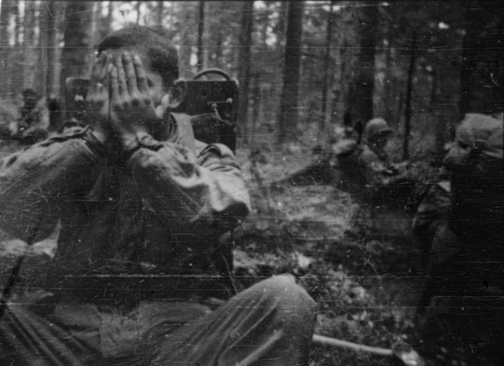 A photo of a soldier in a forested area during the Rescue of the Lost Battalion. The soldier is sitting on the ground holding his face in his hands. Other soldiers are visible in the background.