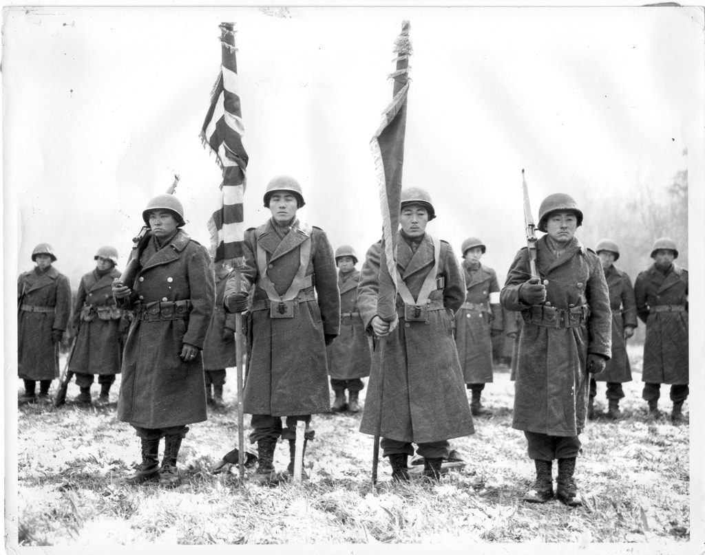 Photo of Japanese American soldiers standing in review in a field in France during WWII. Four soldiers stand in front, two holding flags in the middle and two holding rifles at either side.