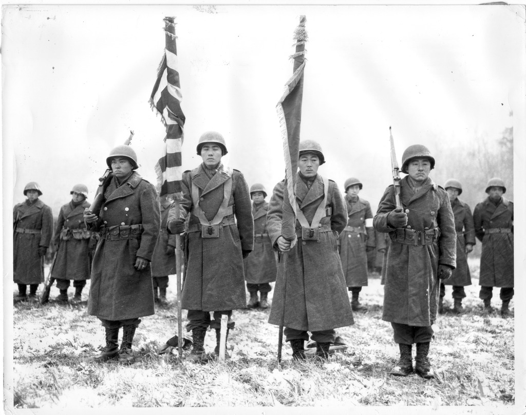 Photo of a parade review of the 442nd following the Bruyères-Biffontaine campaign. The soldiers are helmets and wearing army-issue coats over their uniforms, and stand in two rows facing the camera. In the front row are two color bearers holding flags and two color guards holding rifles over their shoulders. They are standing on a grass field covered in frost.