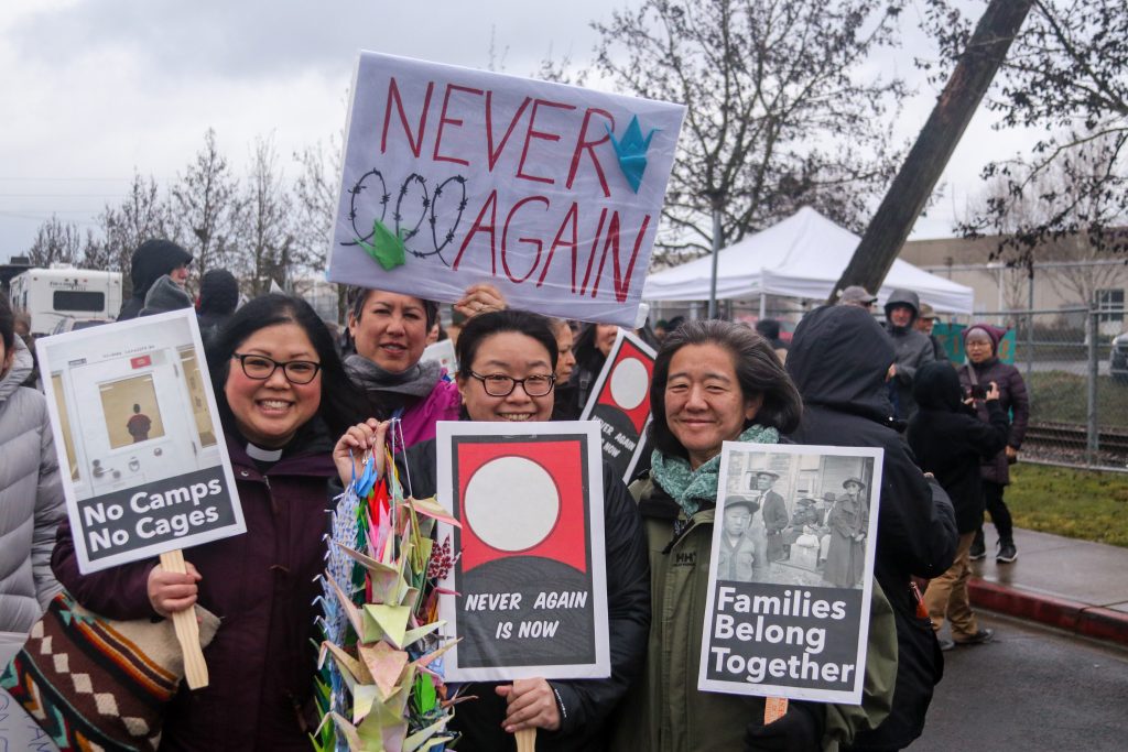 Color photograph of Japanese Americans protesting immigrant detention. They are holding strings of origami cranes and signs that read "No Camps No Cages," "Never Again Is Now," and "Families Belong Together."