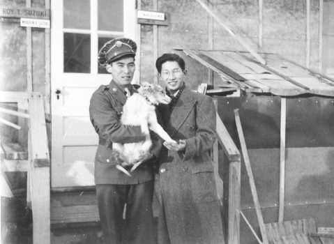 Two men holding a dog in front of a barrack in a Japanese American incarceration camp. The men stand side by side with the dog in the middle. The man on the left wears an army uniform, and the man on the right is wearing glasses and a peacoat. The dog is white/light brown.