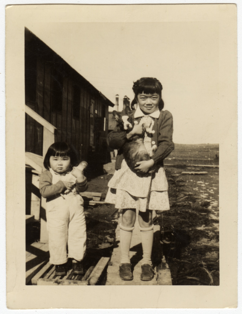 Two young girls stand on the steps in front of a barrack in a Japanese American incarceration camp. The girl at left is a toddler wearing overalls, and the older girl at right is wearing a sweater over a dress and holding puppy.