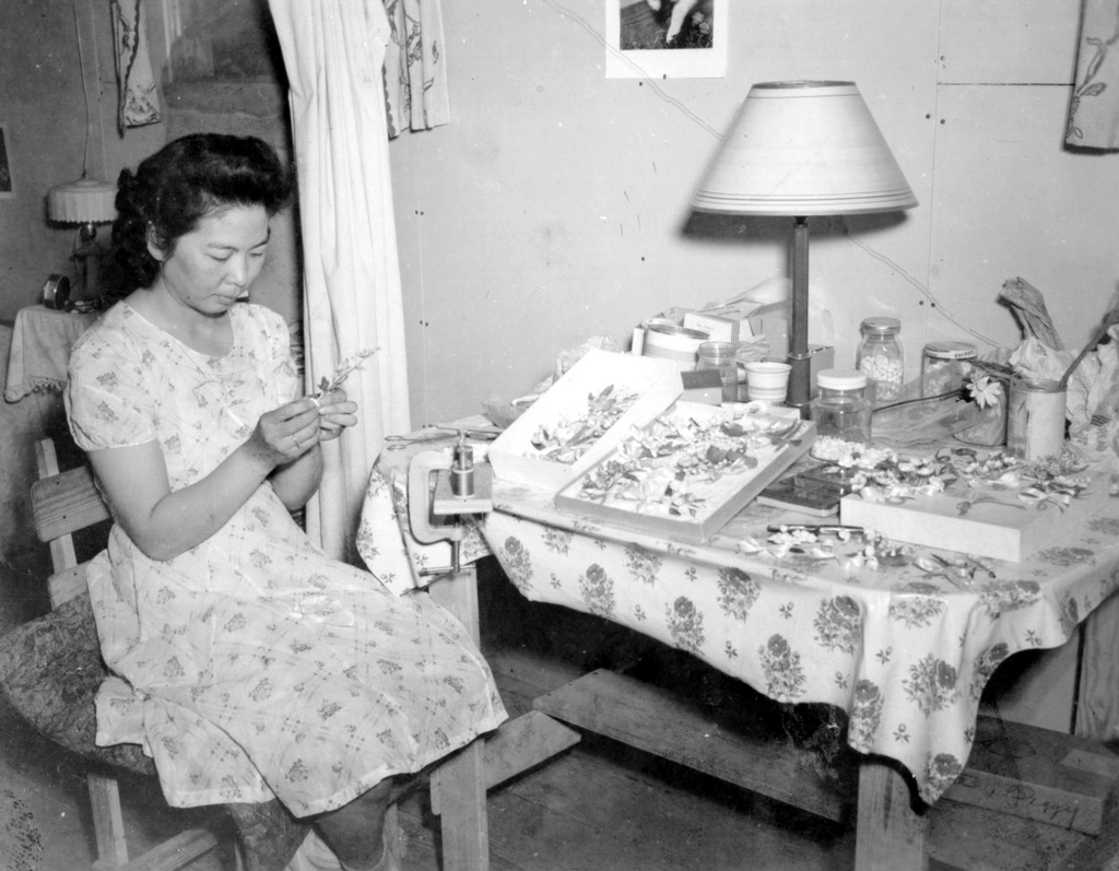A Japanese American woman making jewelry from shells inside a barrack at Tule Lake concentration camp. The woman is wearing a white patterned dress and working on a corsage. She is seated next to a table covered in other pieces of jewelry and crafting supplies.