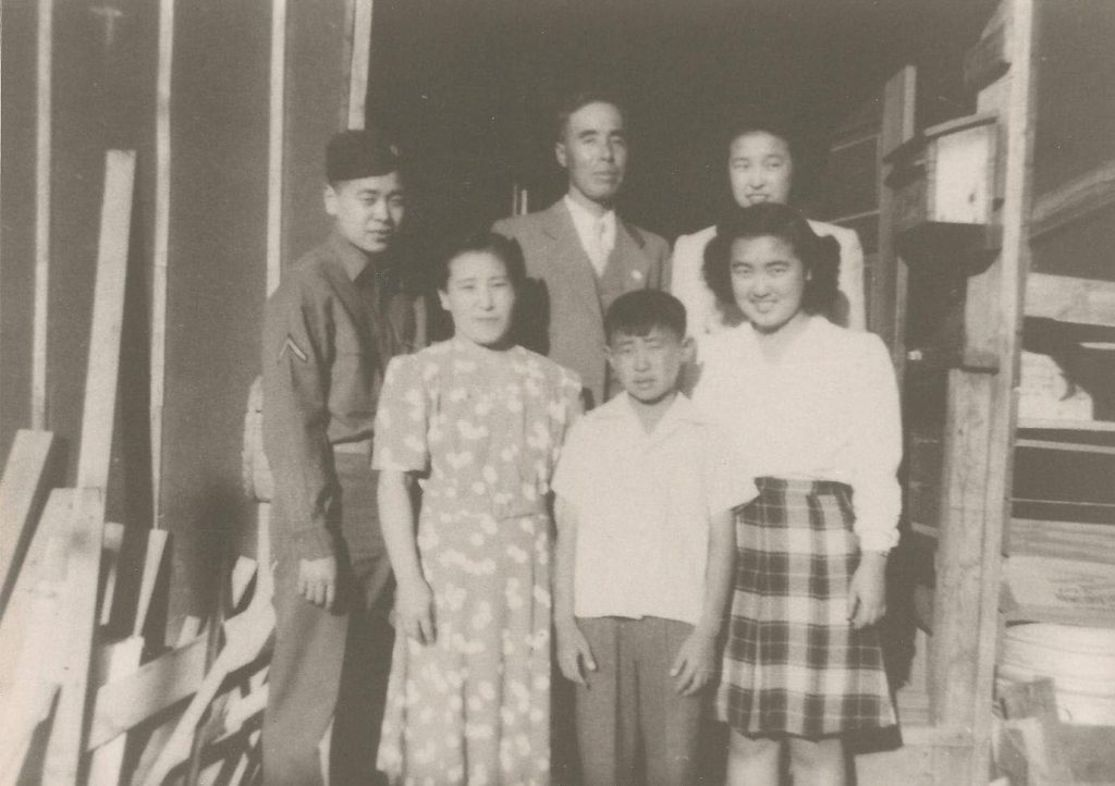 Black and white photo of a Japanese American family in Heart Mountain concentration camp during WWII. The family of six is standing on the steps in front of their barrack in two rows