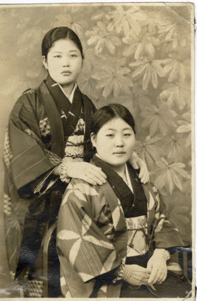 Portrait of two Japanese women wearing kimono and white gloves. One woman is seated in a chair and the other stands behind her resting her hands on her shoulders.