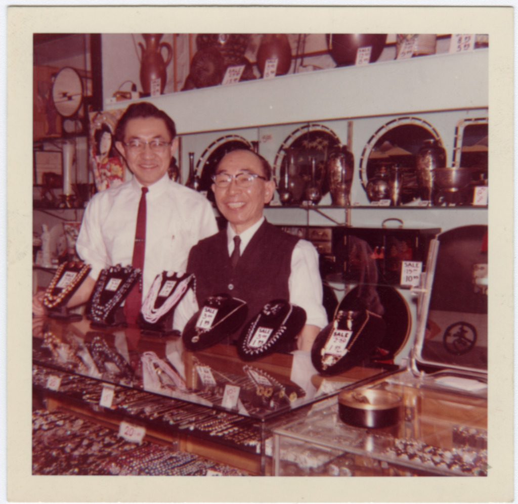 Color photograph of two Japanese American men posing behind the jewelry counter in their gift shop. Both men are wearing glasses and ties and smiling at the camera.
