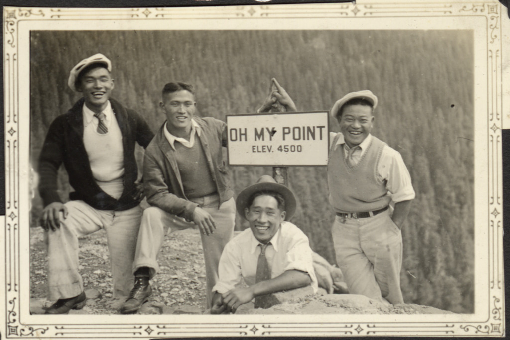 Black and white photo of four Japanese American men on a hiking trip to Mount Rainier National Park. They are smiling and laughing and two of the men are posing with their fingers touching to form a peak over a sign that reads "Oh My Point, elev. 4500."