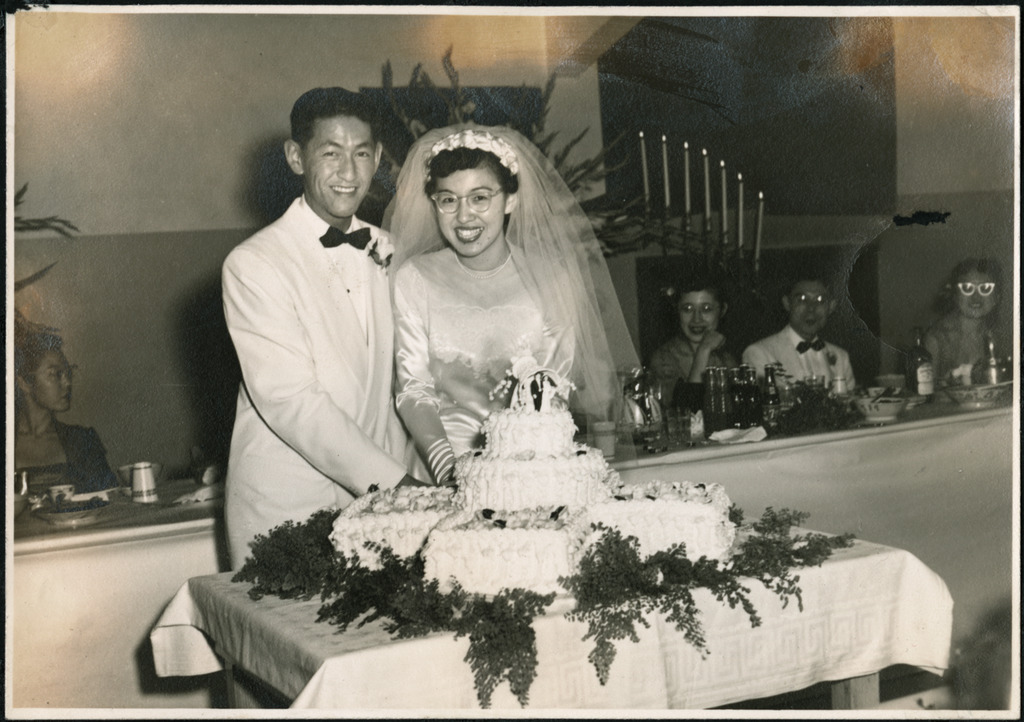 Black and white photo of a Japanese American couple cutting their wedding cake. The bride is wearing a long-sleeved white silk dress and a veil, and the groom is wearing a white suit. Behind them members of the bridal party sit at a banquet table.