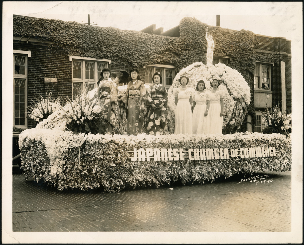 Black and white photo of young women in kimono and white Western dresses stand on a Japanese Chamber of Commerce parade float.