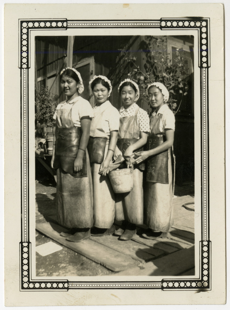 Black and white photo of four Japanese American women cannery workers in pre-war Terminal Island. They are wearing white caps over their hair and rubber aprons.