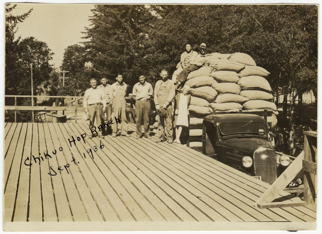 Photo of hops farmers and workers on the Chikuo Hop Ranch in pre-war Oregon. Five men and a girl are standing on a wooden platform next to a truck, and two men are sitting on filled canvas bags loaded onto the truck bed. A handwritten note on the front of the photo reads "Chikuo Hop Ranch Sept. 1936"