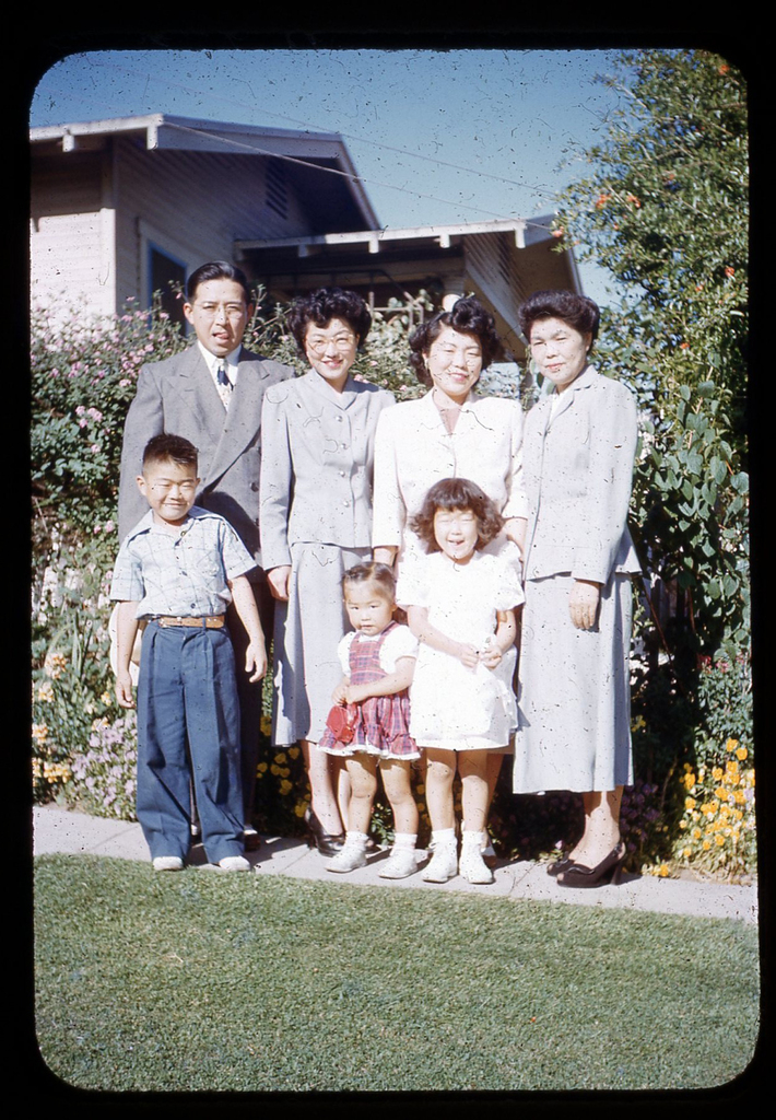 Color photo of a Japanese American family posing in front of a house. A man and three women stand in a row behind three children.