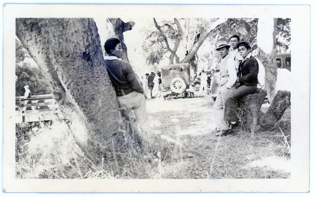Black and white photo of four men lounging outside. One man leans against a tree on the left and three others are sitting or standing on the right. A car and other people are visible in the background.