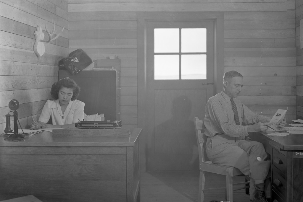 Black and white photo of an administrative office in the Minidoka concentration camp during WWII. A Japanese American woman is working at a desk on the left, and a white man is seated at another desk on the right reading a pamphlet.