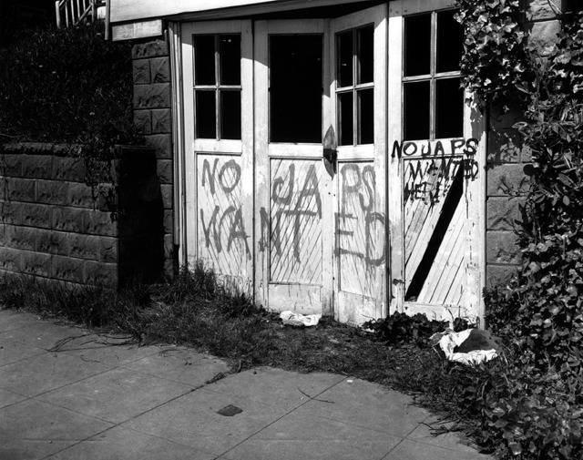 Black and white photo showing racial slurs spray painted on the garage door of a Japanese American family. The door is white, with four-paned windows. The graffiti reads "No Japs Wanted" and "No Japs Wanted Here."