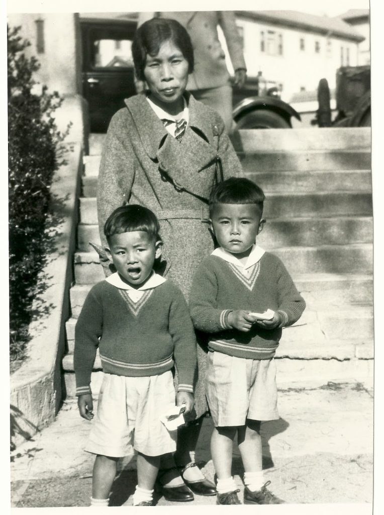 Black and white photo of a Japanese American and two young boys posing in front of a set of concrete steps. The woman is wearing what looks like a wool coat, and the boys are wearing matching shorts and sweaters.