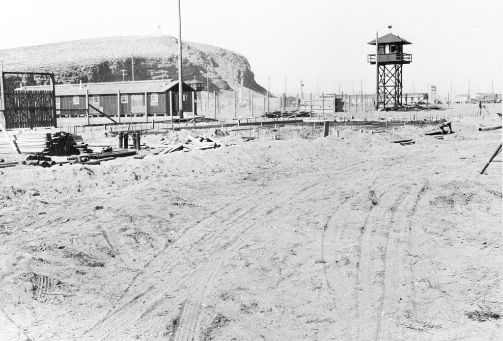 Photo of Tule Lake concentration camp during WWII. Guard towers and barracks buildings are visible in the background, behind a fence. There are wood planks lying on a construction site in the foreground.