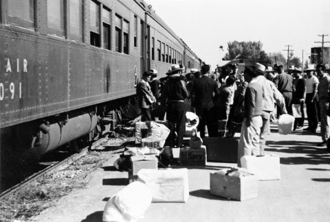 Photo showing Japanese Americans waiting to board a train to a WWII concentration camp. A crowd is standing next to a train, with their luggage on the ground.
