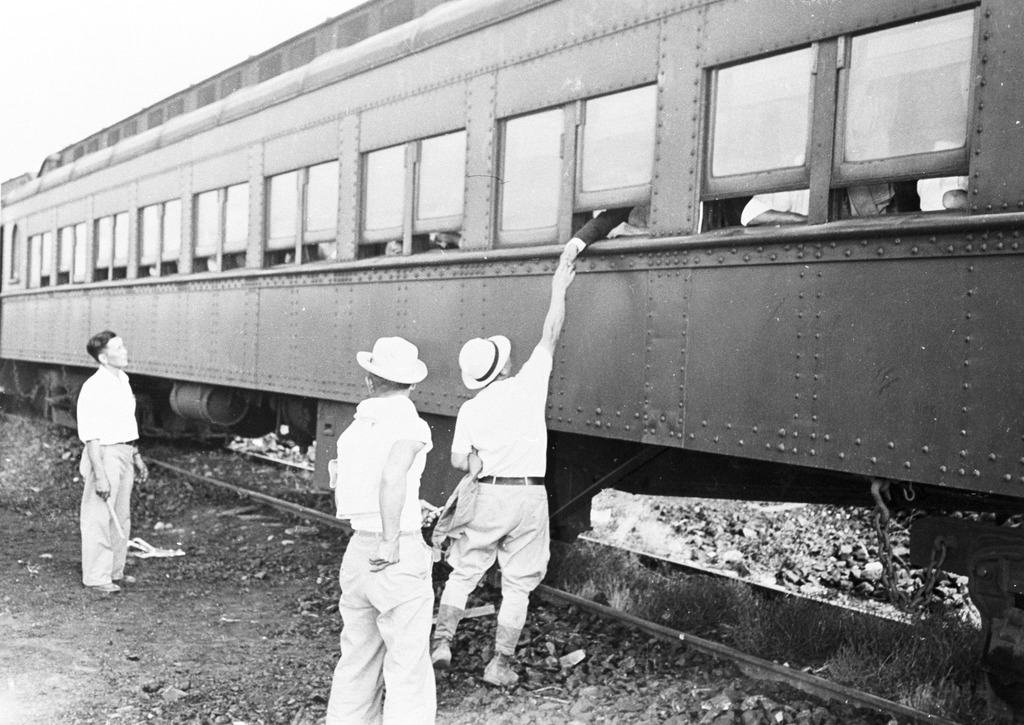 Photo of Japanese Americans saying goodbye to friends being transferred to another concentration camp during WWII. Three men are standing next to the train tracks, one of them shaking the hand of someone reaching out of the train window.