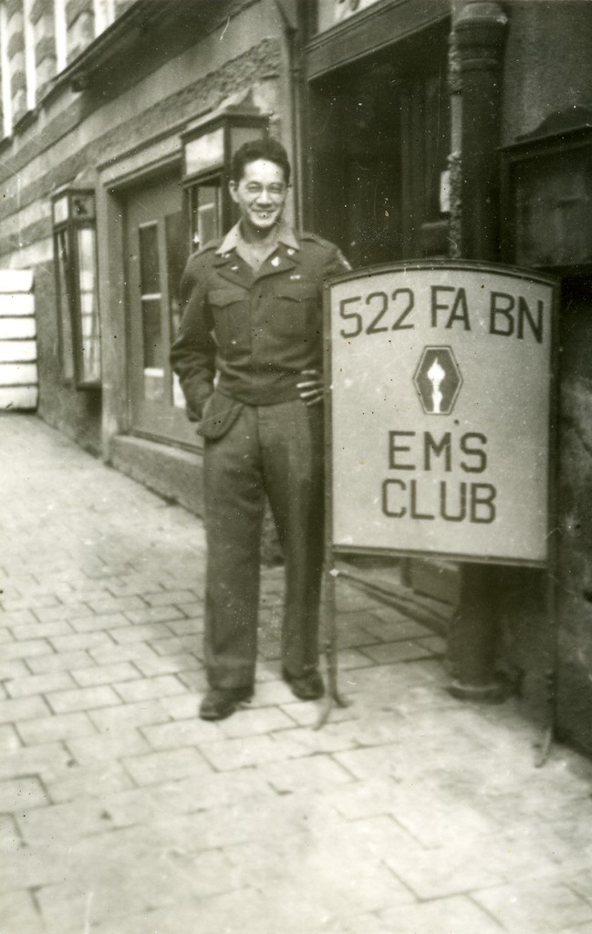 A Japanese American soldier posing in uniform next to a sign that reads "522 FA BN EMS Club"