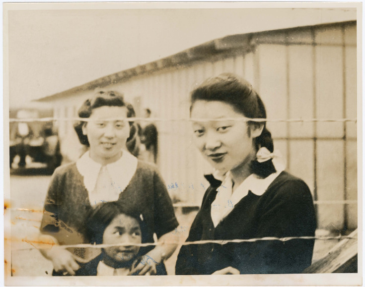 Black and white photo of two Japanese American women and a girl in a WWII concentration camp. Both women are wearing dark sweaters over light blouses and looking at the camera from behind a barbed wire fence. The woman at left stands behind the girl, resting her hands on the girl's shoulders. Visible in the background is a barracks and a car.