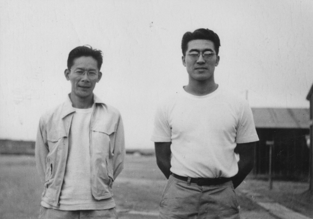 Two leaders of the Heart Mountain Fair Play Committee standing outside a barrack. Both men are standing with their arms resting behind their backs, looking at the camera.
