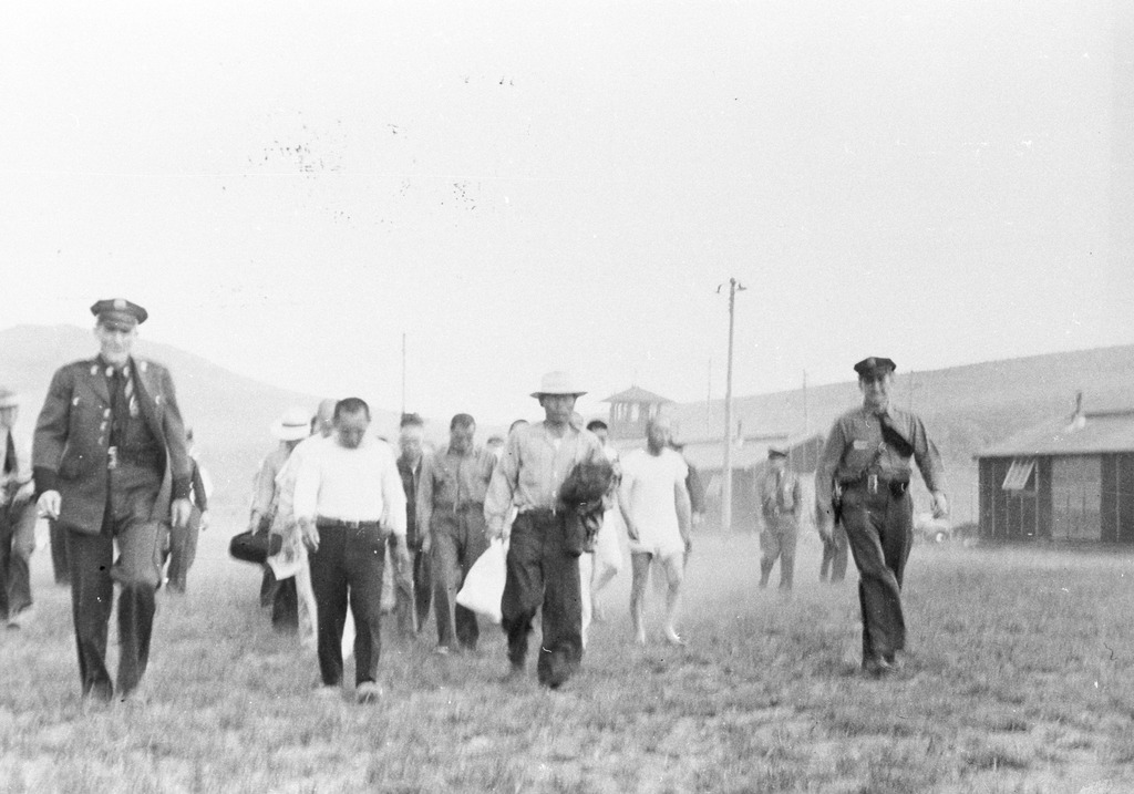 Somewhat blurry black and white photo of Japanese American men being escorted through Tule Lake by military police. Several of the men are carrying bags. One is wearing only a t-shirt and boxers and appears to be barefoot. A guard tower and two barracks are visible in the background.