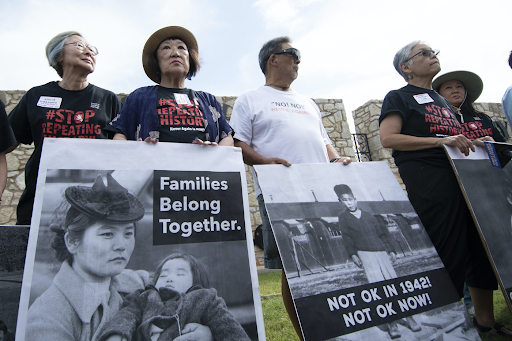 Color photo of four Japanese American survivors of WWII incarceration protesting the detention of immigrants at Fort Sill in June 2019. They are wearing t-shirts that read "#StopRepeatingHistory" and holding signs with pictures from Japanese American incarceration that read "Families Belong Together" and "Not OK in 1942! Not OK now!"