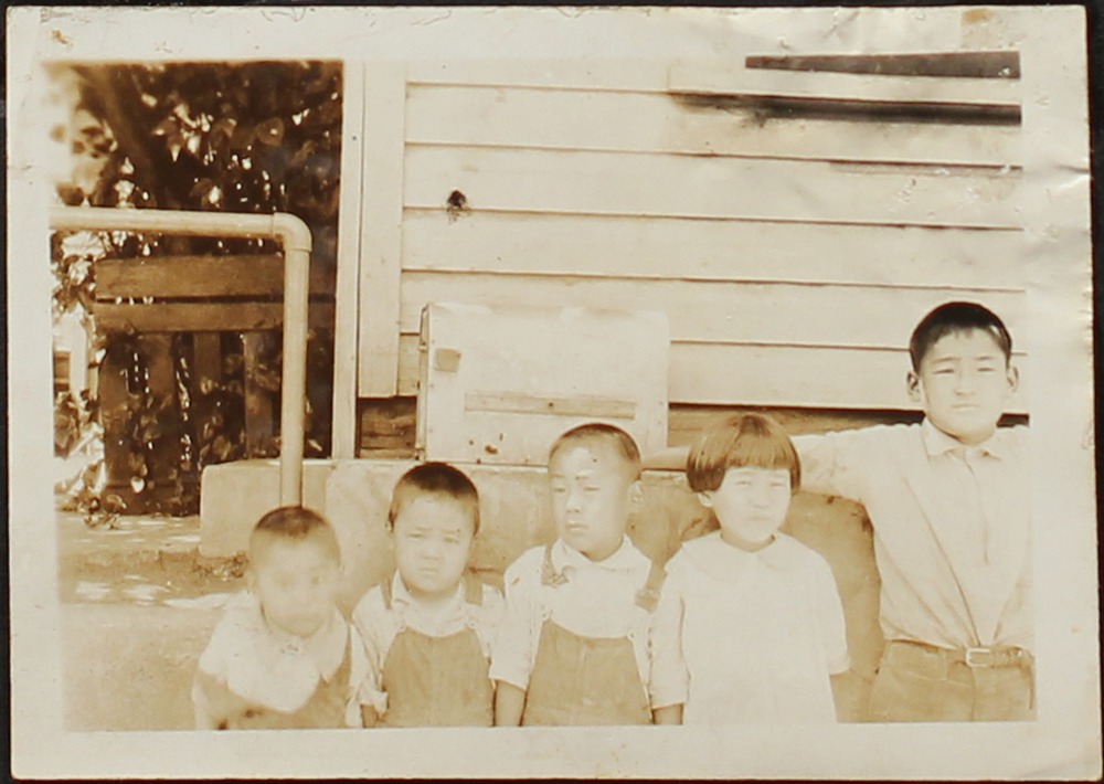 Black and white photo of five Japanese American children in front of a house. They are standing in a line from youngest to oldest.