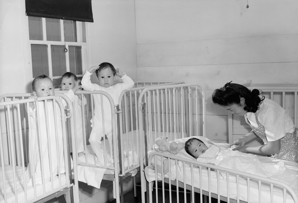 Black and white photo of the orphanage in Manzanar. Three toddlers are standing up in white cribs next to a window, while a nurse tucks another toddler into bed.