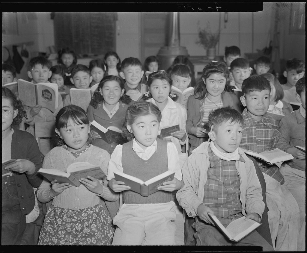 Black and white photo of Japanese American schoolchildren inside a concentration camp classroom. The children are holding books open in their laps and looking off camera, presumable at their teacher.