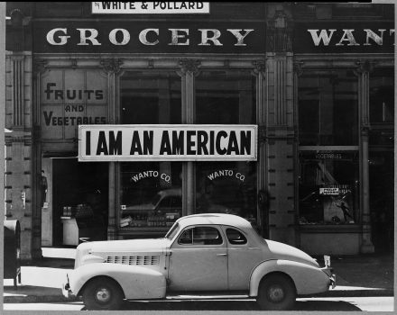 Black and white photo of a grocery storefront. The words "Fruits and Vegetables" and "Wanto Co." are painted on the windows, and a large white sign with black lettering that reads "I AM AN AMERICAN" is posted on the building. A car is parked on the street in front of the store.