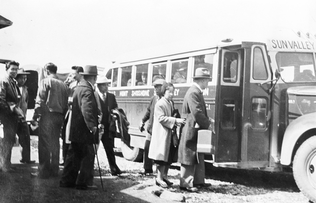 Black and white photo of Japanese Americans boarding a bus at Minidoka. Bus says Sun Valley in front and the words Hunt and Shoshone can be read on the side of the bus. A man and a woman are preparing to board and some young men are milling about. An army truck is seen in the background.