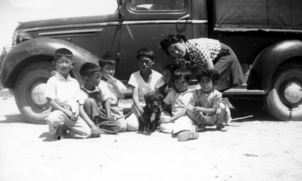 Black and white photo of a Japanese American woman and seven children posing with a dog in front of a black truck.