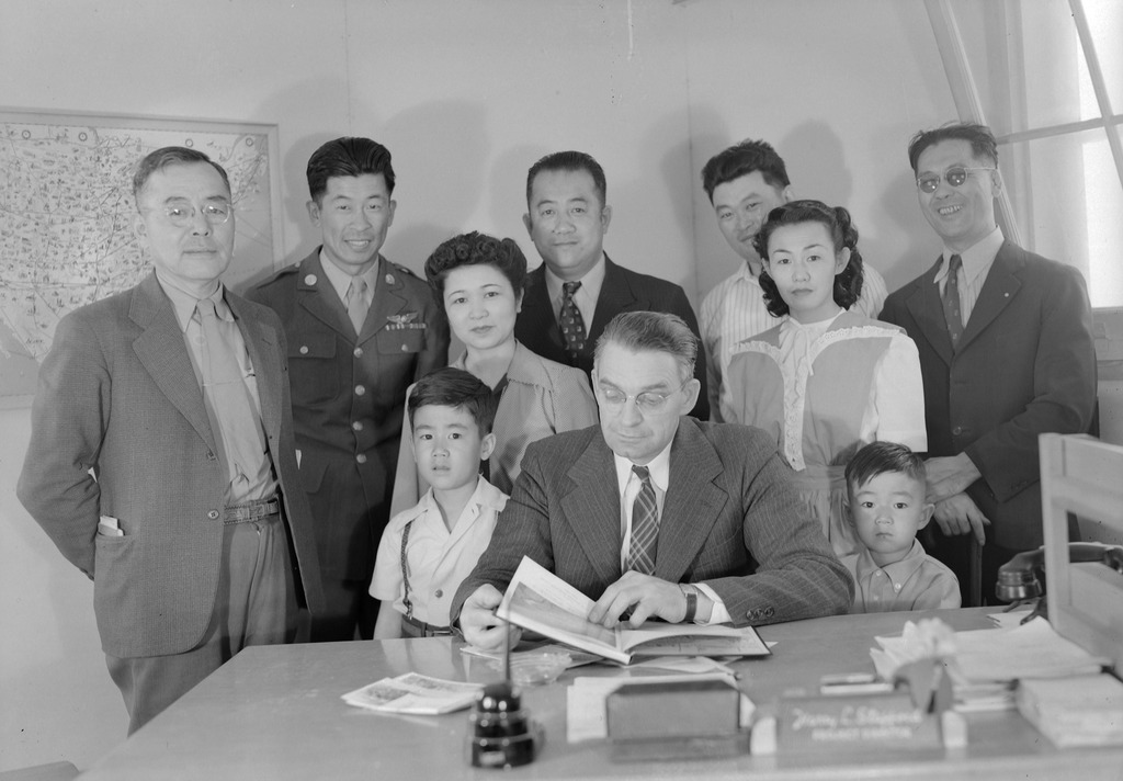 Black and white group photo of seven Japanese American men and women and two children with the "project director" of a WWII concentration camp. The director is seated at his desk in the center.