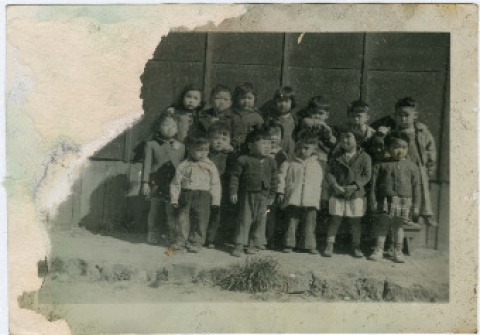 Black and white photo of elementary school children standing outside a barrack in a Japanese American concentration camp. The left upper corner of the photo is torn.