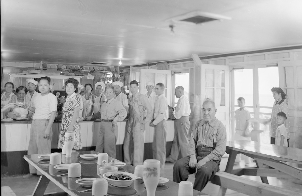Black and white photo of Japanese Americans in the mess hall of a WWII concentration camp. Workers wearing aprons stand behind a counter in the background serving food to people standing in a line. There are two tables in the foreground, one set with plates and utensils. An elderly man is seated at an empty table near two open doors.