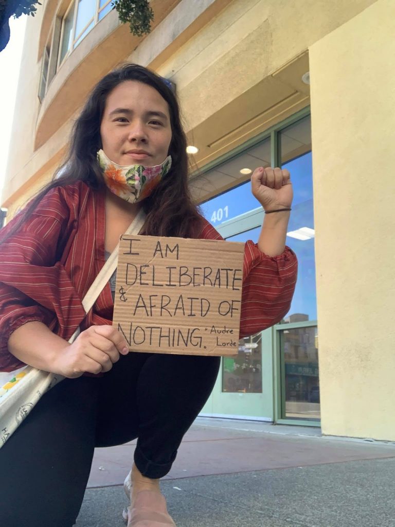 Photo of a Nikkei person with long hair and a floral face mask pulled down over their chin, kneeling on the sidewalk in front of a yellow building. They are posing with a raised fist and a cardboard sign that reads, "I am deliberate & afraid of nothing. -Audre Lorde"
