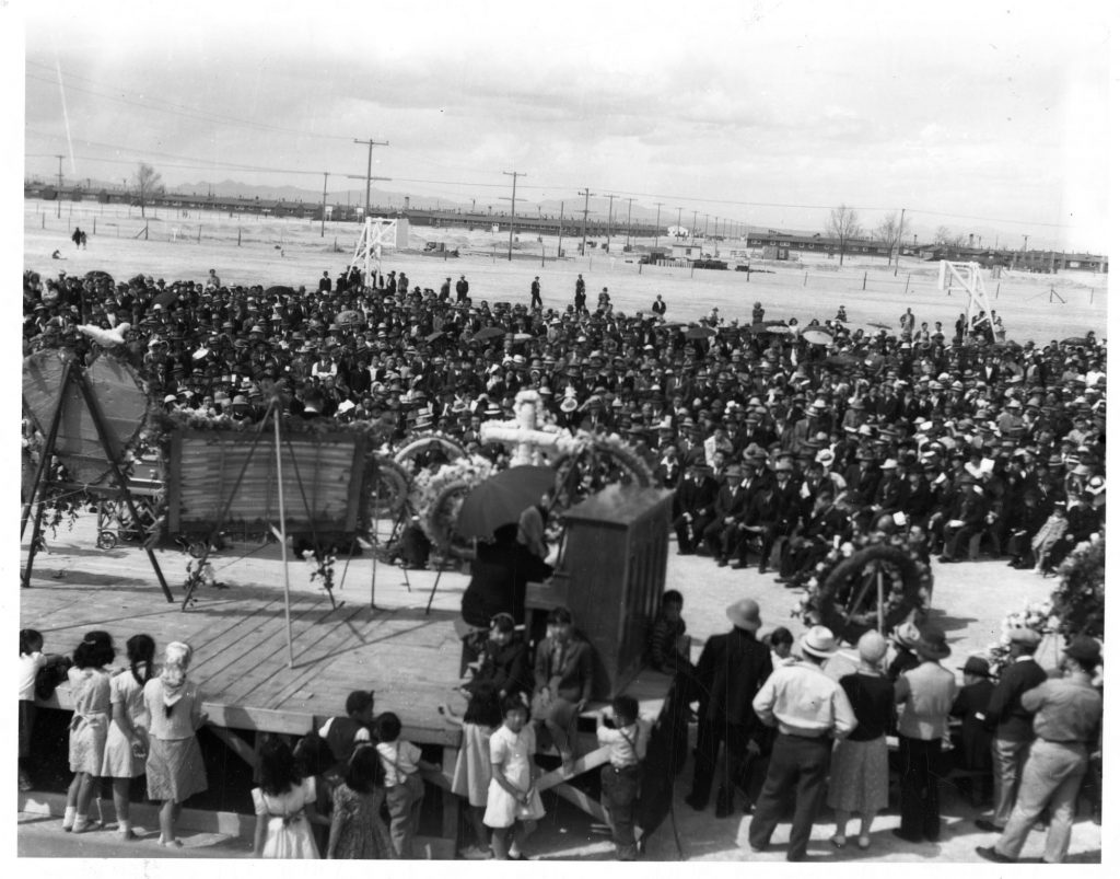 Black and white photo of a funeral in Topaz concentration camp. Hundreds of mourners are seated or standing in front of a wooden stage with funeral wreaths and a piano. The photo was taken from behind the stage, where additional mourners and standing to watch. Barracks buildings are visible in the distance.