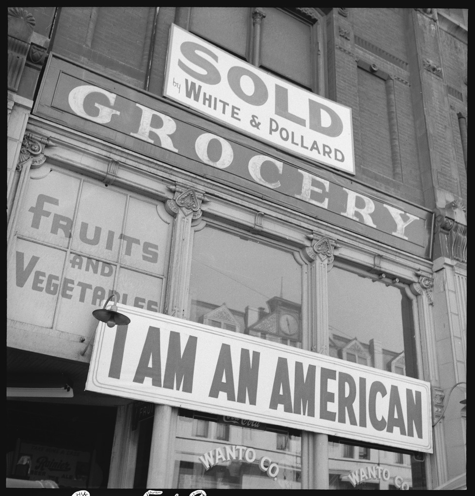 Black and white photo of a grocery storefront with the words "Fruits and Vegetables" and "Wanto Co." painted on the windows. There are two large white signs with black lettering posted on the storefront. One reads, "SOLD by White & Pollard" and the other reads, "I AM AN AMERICAN."