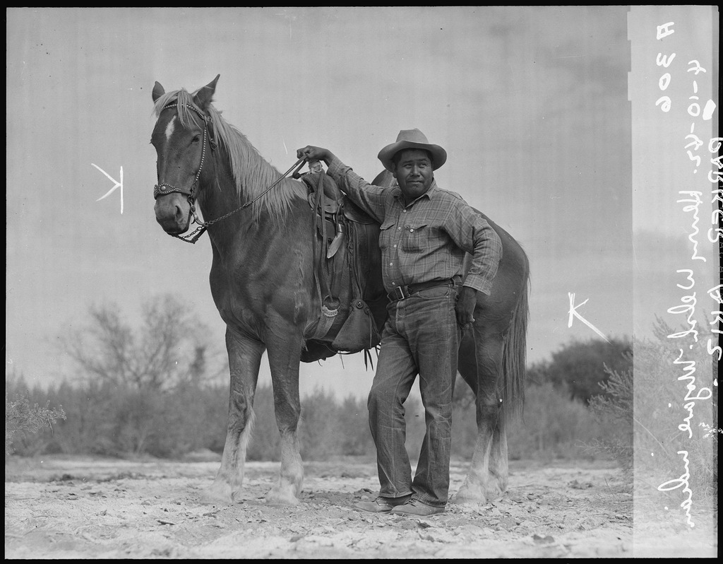Photo of Henry Walsh, a member of the Mohave Tribe and chairman of the Colorado River Indian Community Tribal Council, taken at Poston concentration camp in 1942. Henry is standing in front of a horse, resting his arm on the saddle.