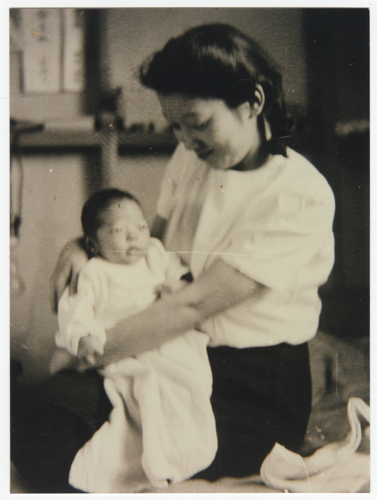 Black and white photo of a young Japanese American woman sitting on what appears to be a bed in a WWII concentration camp, holding her infant son.