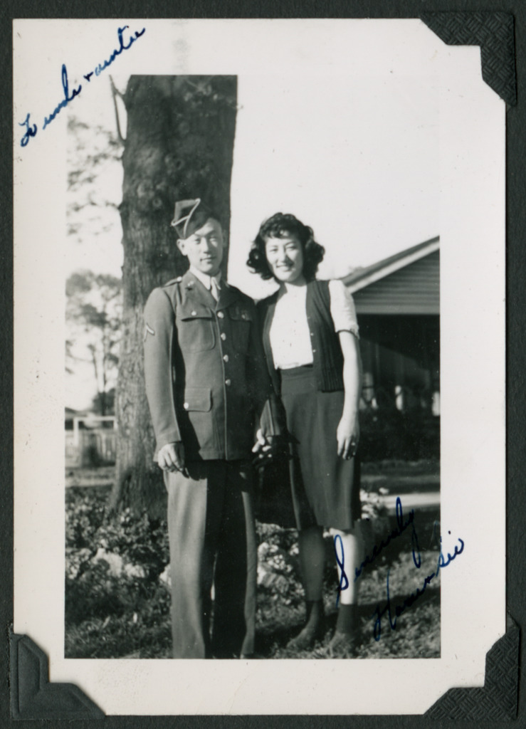 Black and white photo of a uniformed Nisei soldier and Japanese American woman holding hands. They stand in front of a tree trunk and a house is visible in the background. In the upper right corner is a handwritten note that reads, "To uncle & auntie."